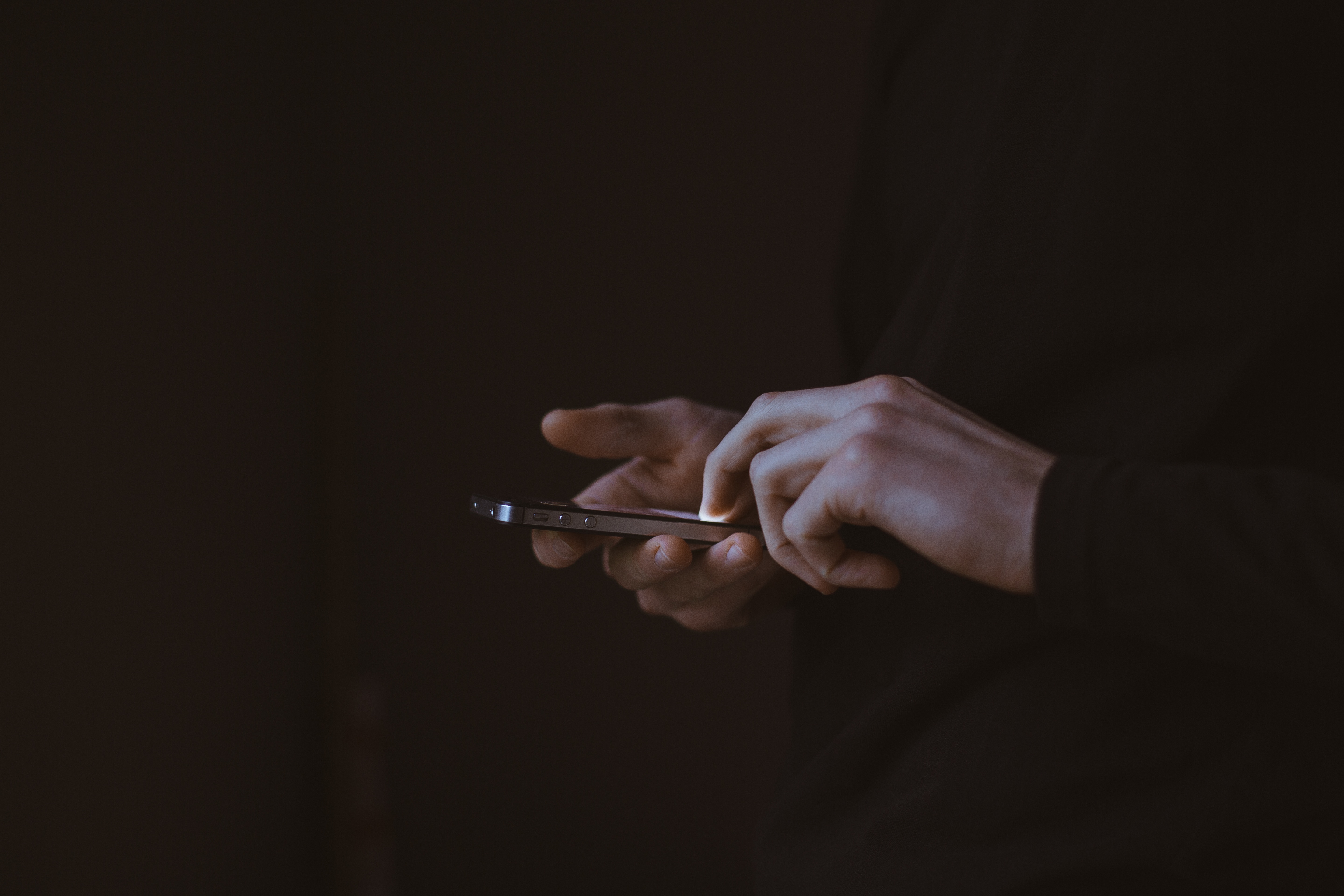 Close up of hand holding and texting on an iPhone. Focus of photo is on hands and phone against an all-black background.