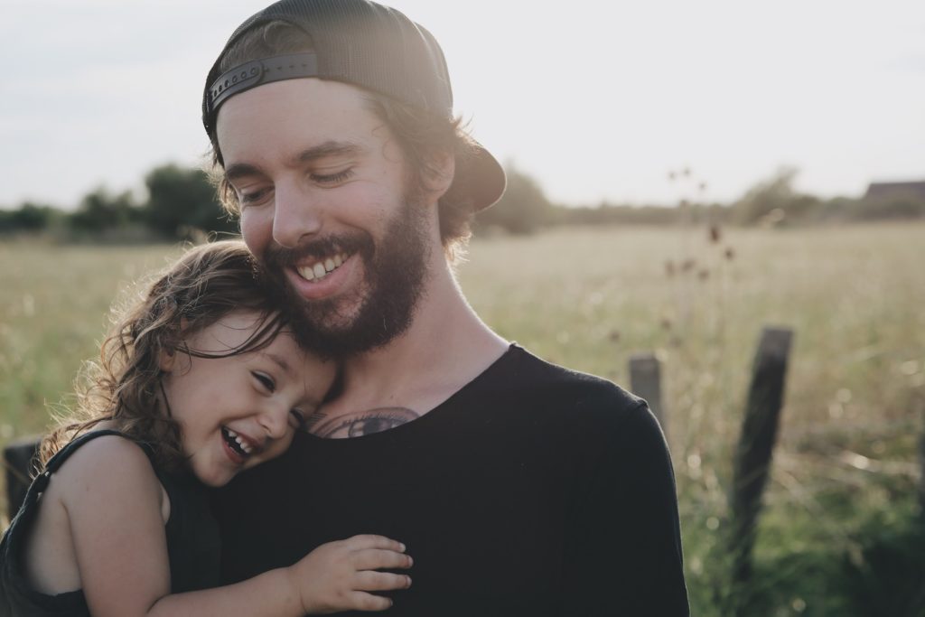 Smiling father with ball cap on backwards holding smiling young daughter, standing in golden field of grass