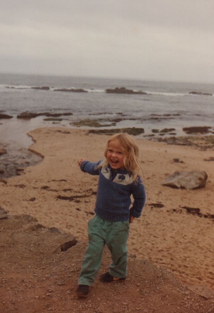 a very happy three year old zoe on a beach pointing at ocean behind her 