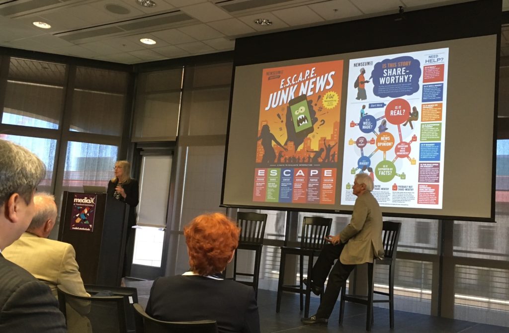 Esther Wojicki at podium in front of screen with ESCAPE Junk News poster