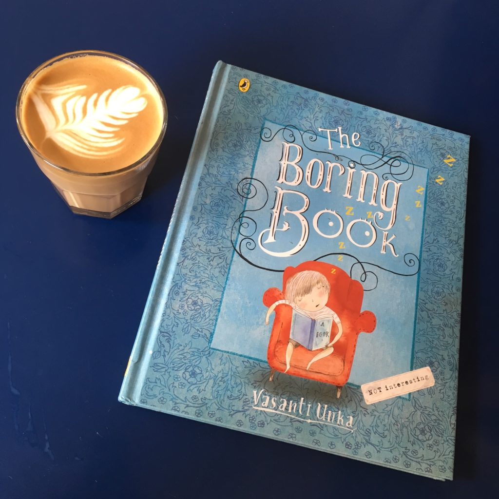 Book titled The Boring Book on cafe table with a coffee drink. On book cover, child sits in big chair falling asleep with book in hands