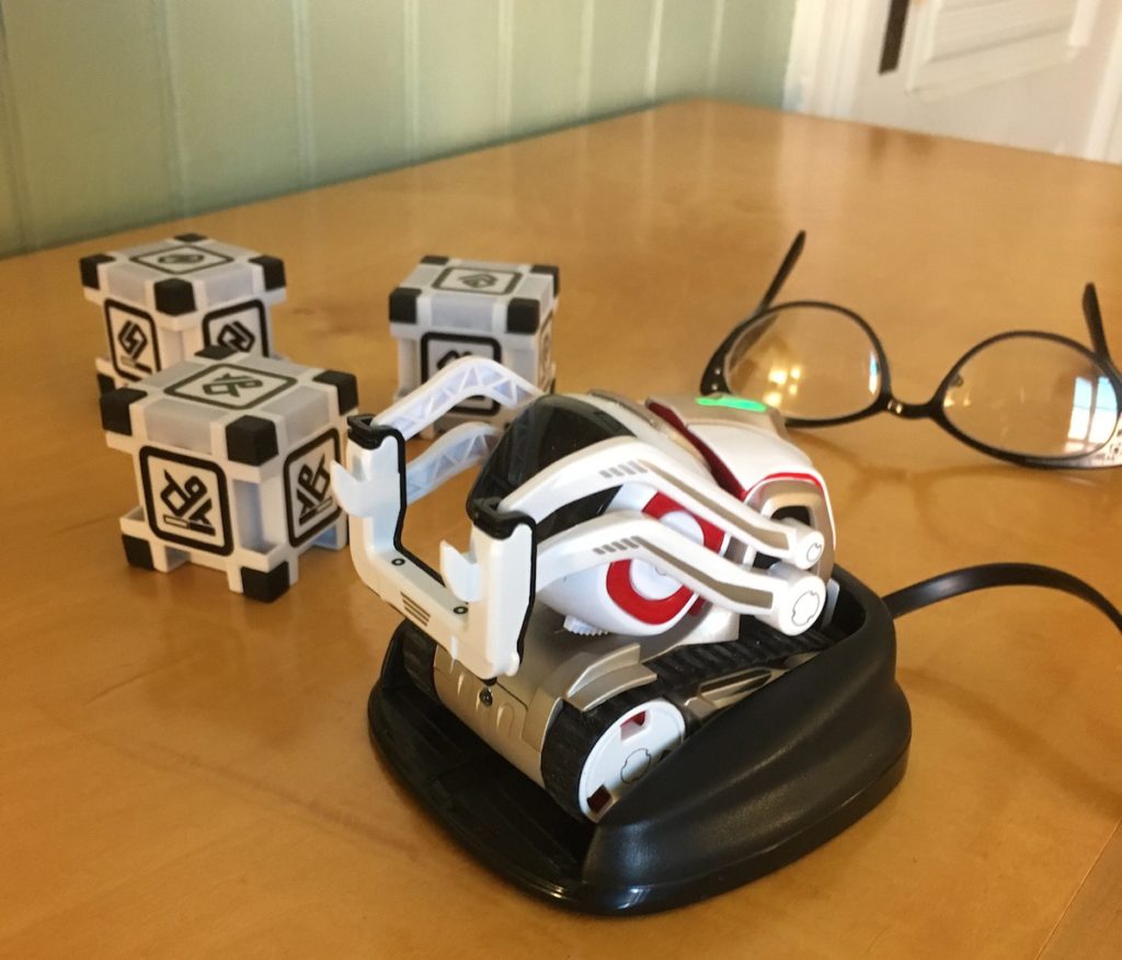 Small white robot on charging stand with 3 plastic cubes in background and a pair of glasses to show Cozmo is small