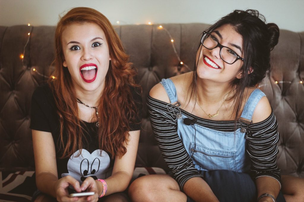 Two young happy women sitting side by side on a couch. One hold a mobile phone in hands