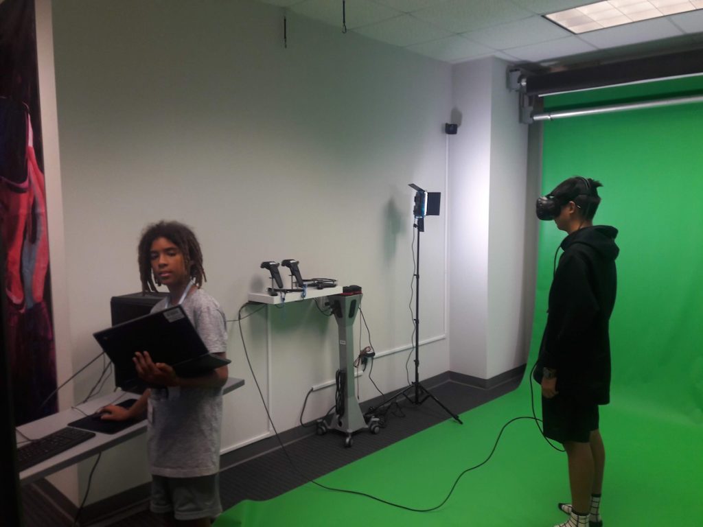 one student with VR headset standing in green room and second student holding laptop on one hand and playing keyboard with other hand