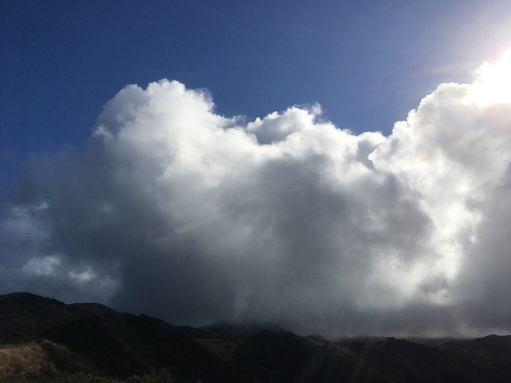 Very large and close rain cloud in blue sky above hills with sun flares behind cloud